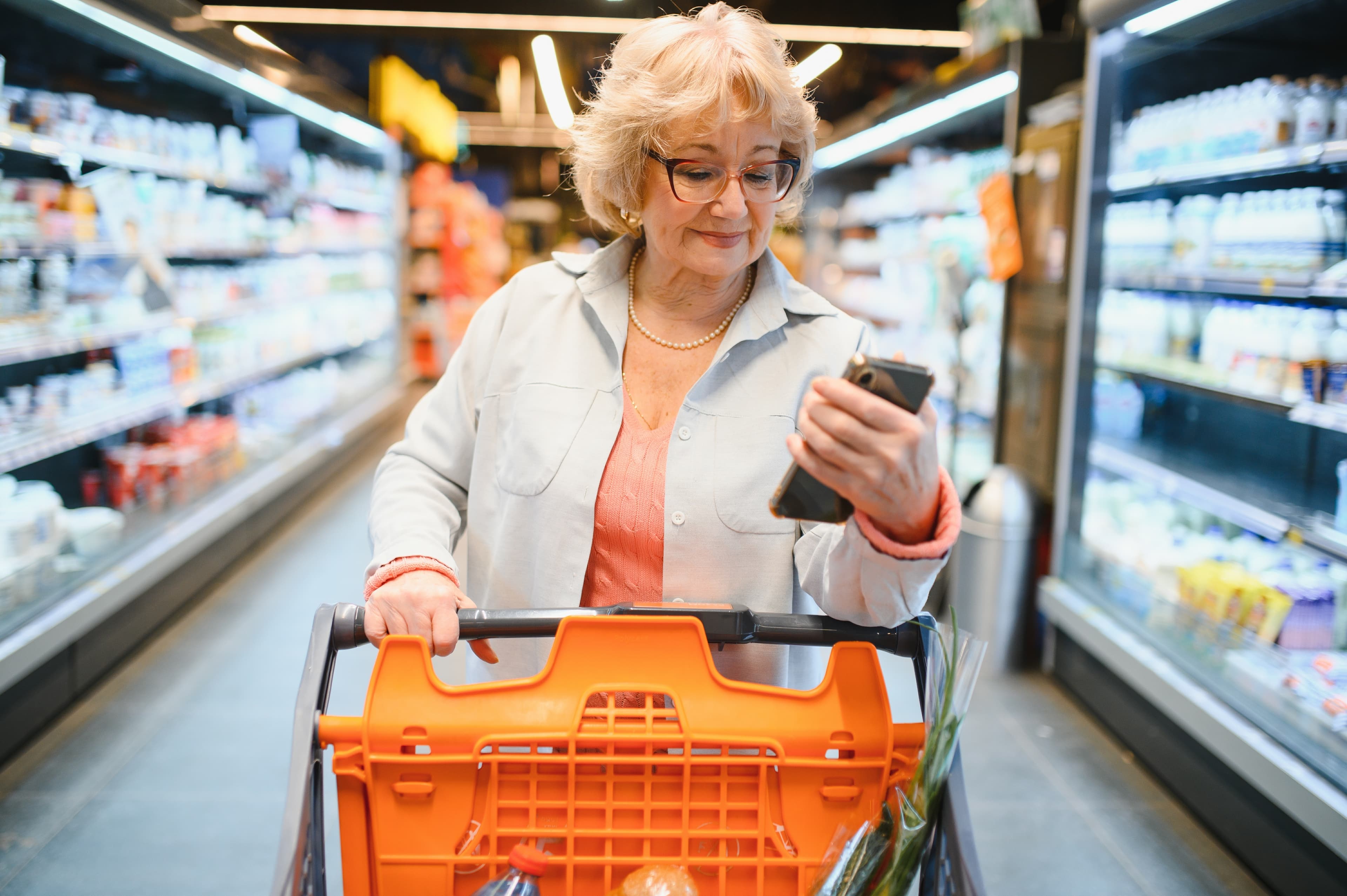 Happy senior woman looking at grocery store aisle with cellphone in hand