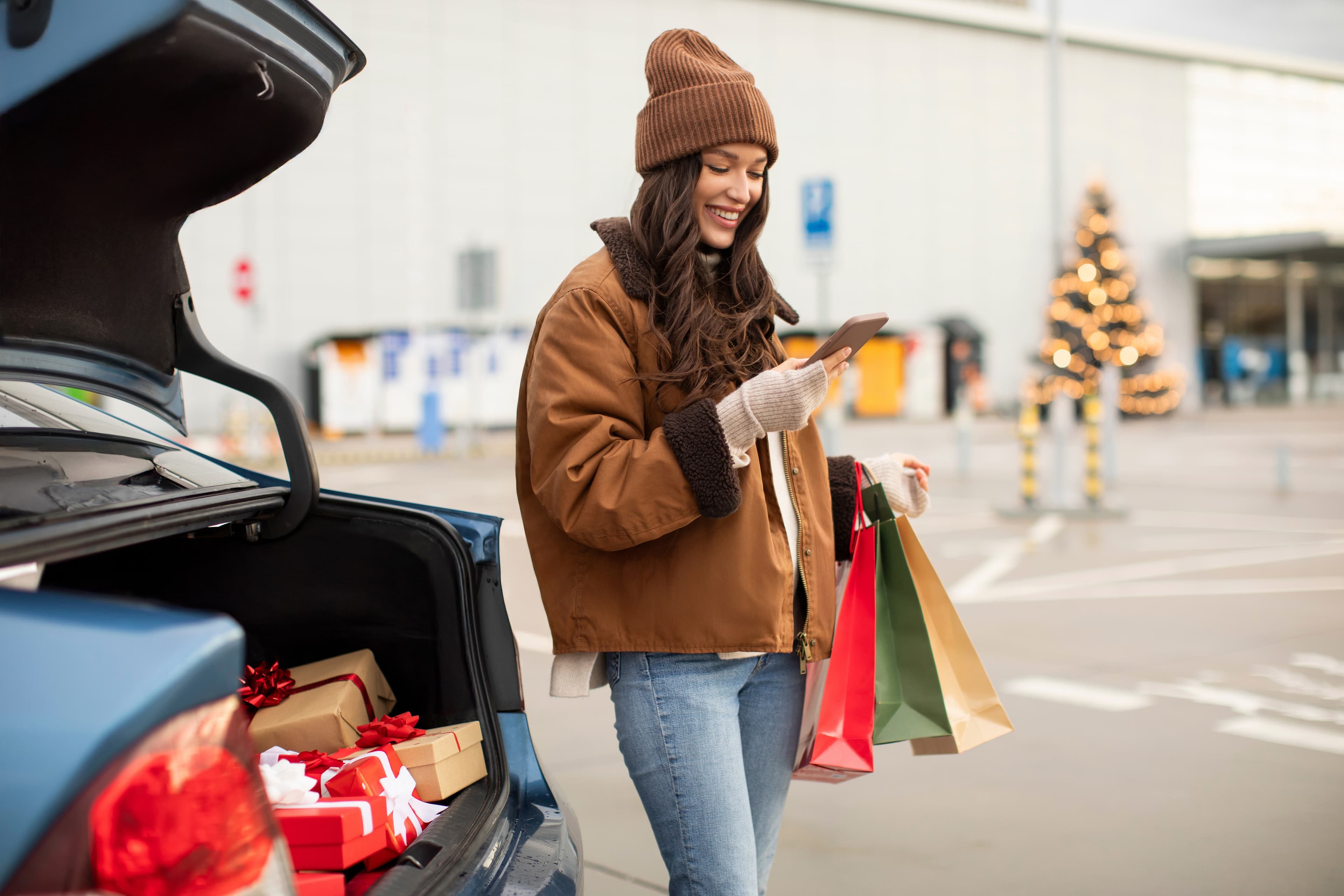 New year mood and shopping concept. Happy woman sits with cellphone and chopper bags standing near car trunk full of Christmas presents near mall outdoors.
